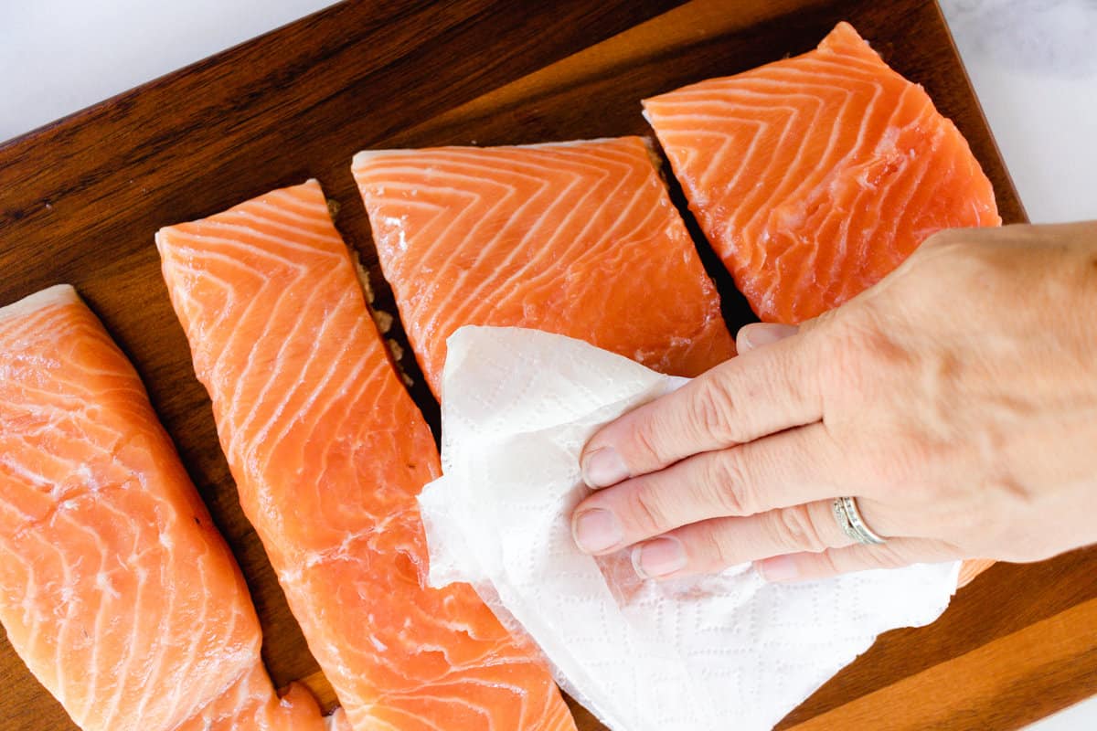 Gently patting the excess moisture off the salmon fillets that are sitting on a wooden cutting board. 