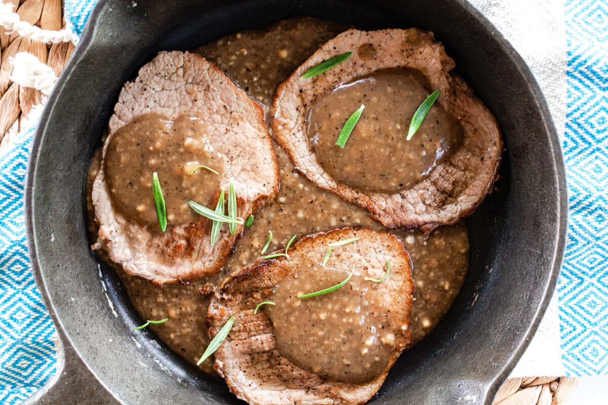 Three cooked pieces of steak in a skillet, drenched with dark gravy and sprinkled with fresh rosemary leaves.