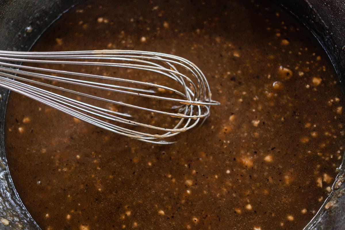 In a dark colored pot is gravy with a whisk leaning against the side.