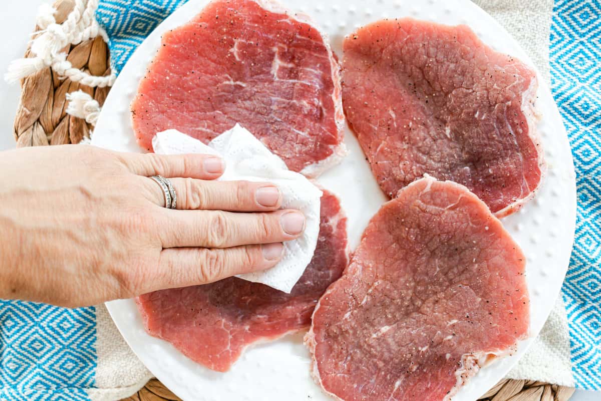 Hand drying eye of round steak that is set on a glass plate on a blue and white table cloth.