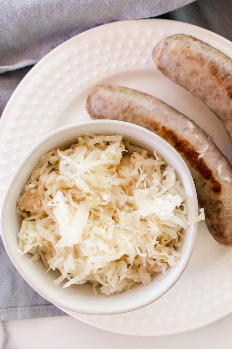 Platted on a round glass plate is two brats and a bowl of fried sauerkraut.