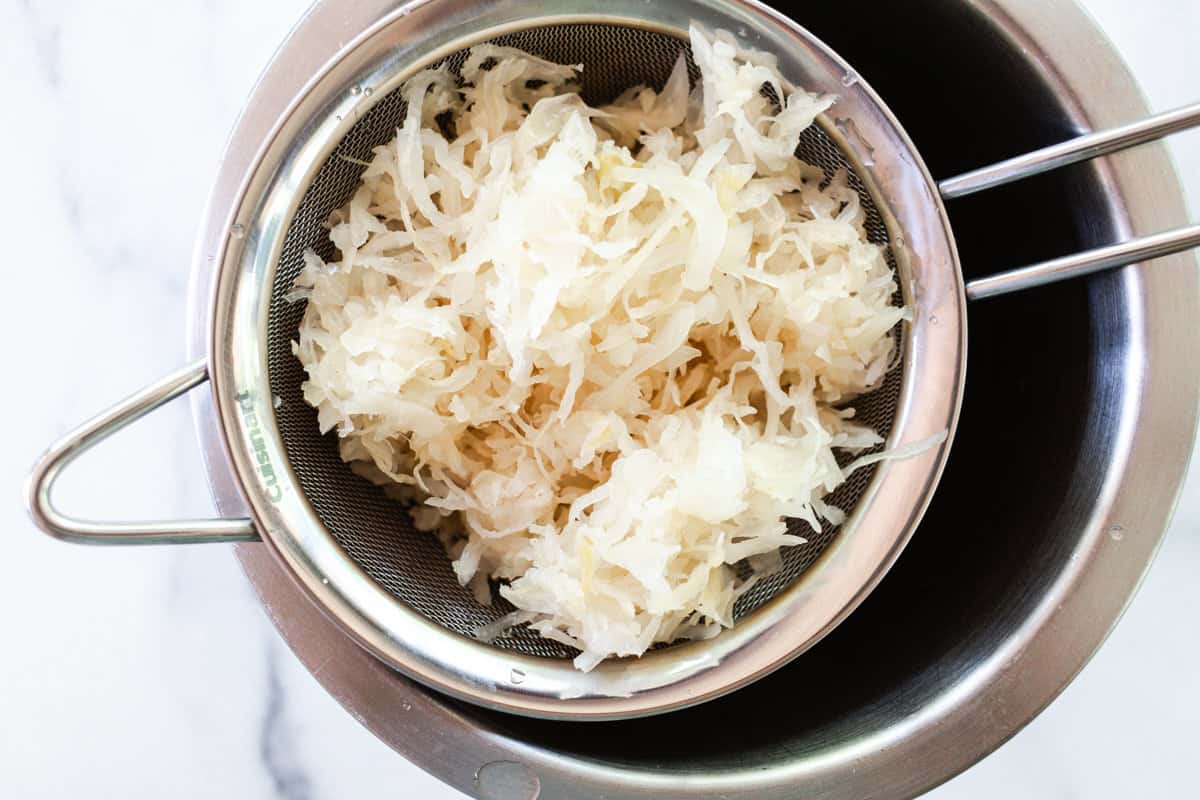 A metal strainer filled with fermented cabbage sitting over a silver bowl.