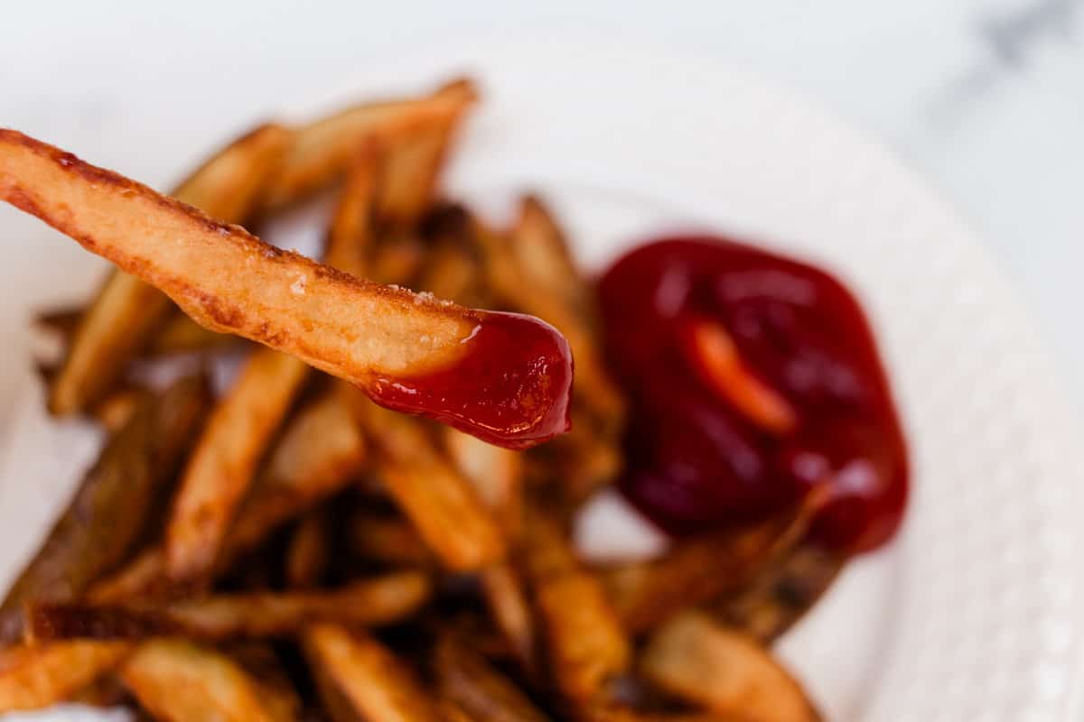 A crispy french fry dipped in ketchup with a plate a fries and sauce in the background.