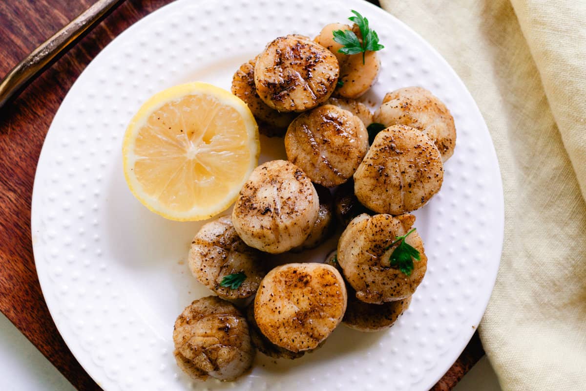 Clean white plate sitting on a wooden board is topped with scallops and a slice of citrus fruit. 