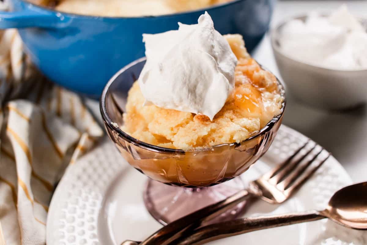 A single serving dessert dish filled with peach dump cake sitting on a glass plate with a fork and spoon on the side. 