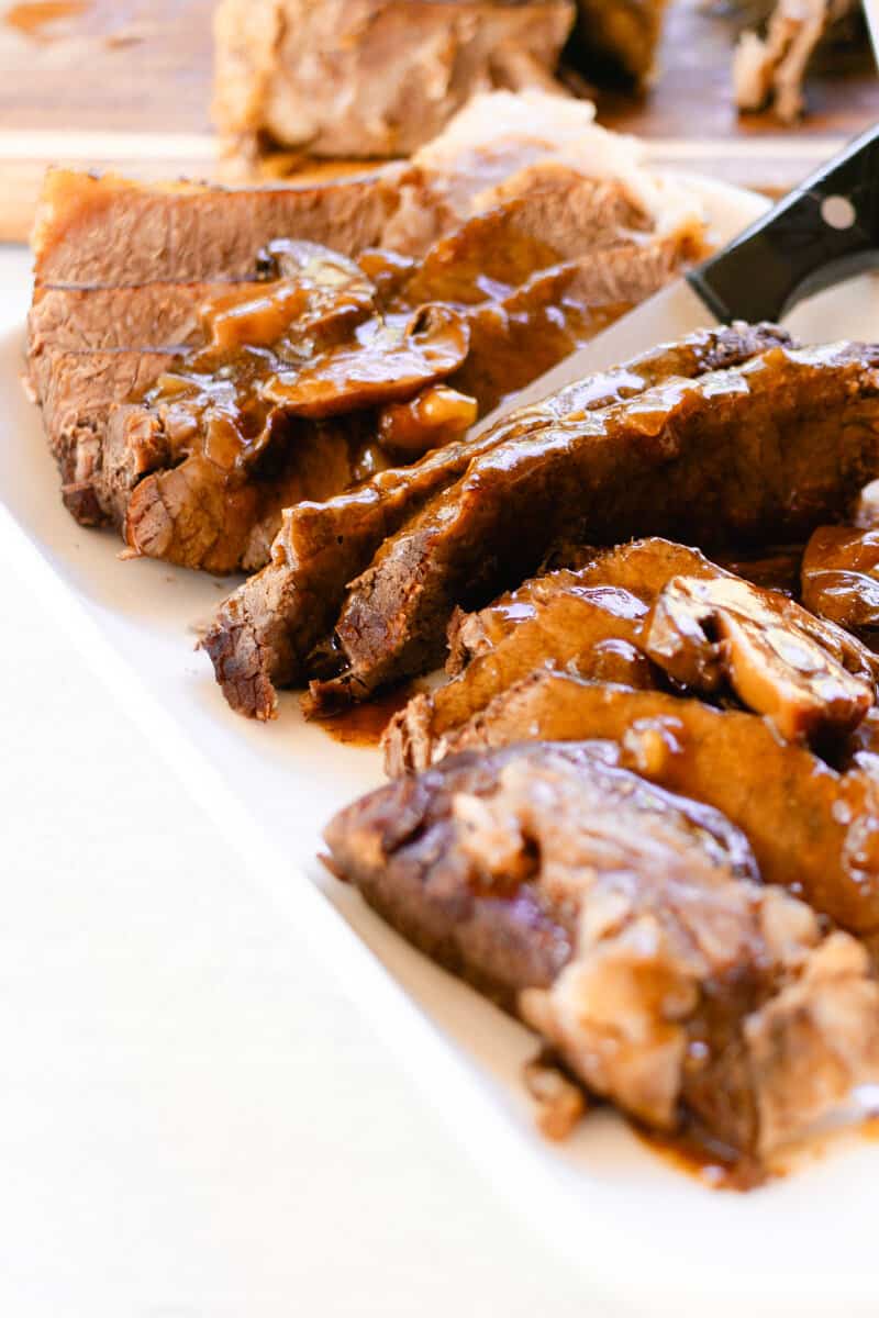 Slicing a pikes peak roast with a sharp knife into thin slices on a white serving plate.