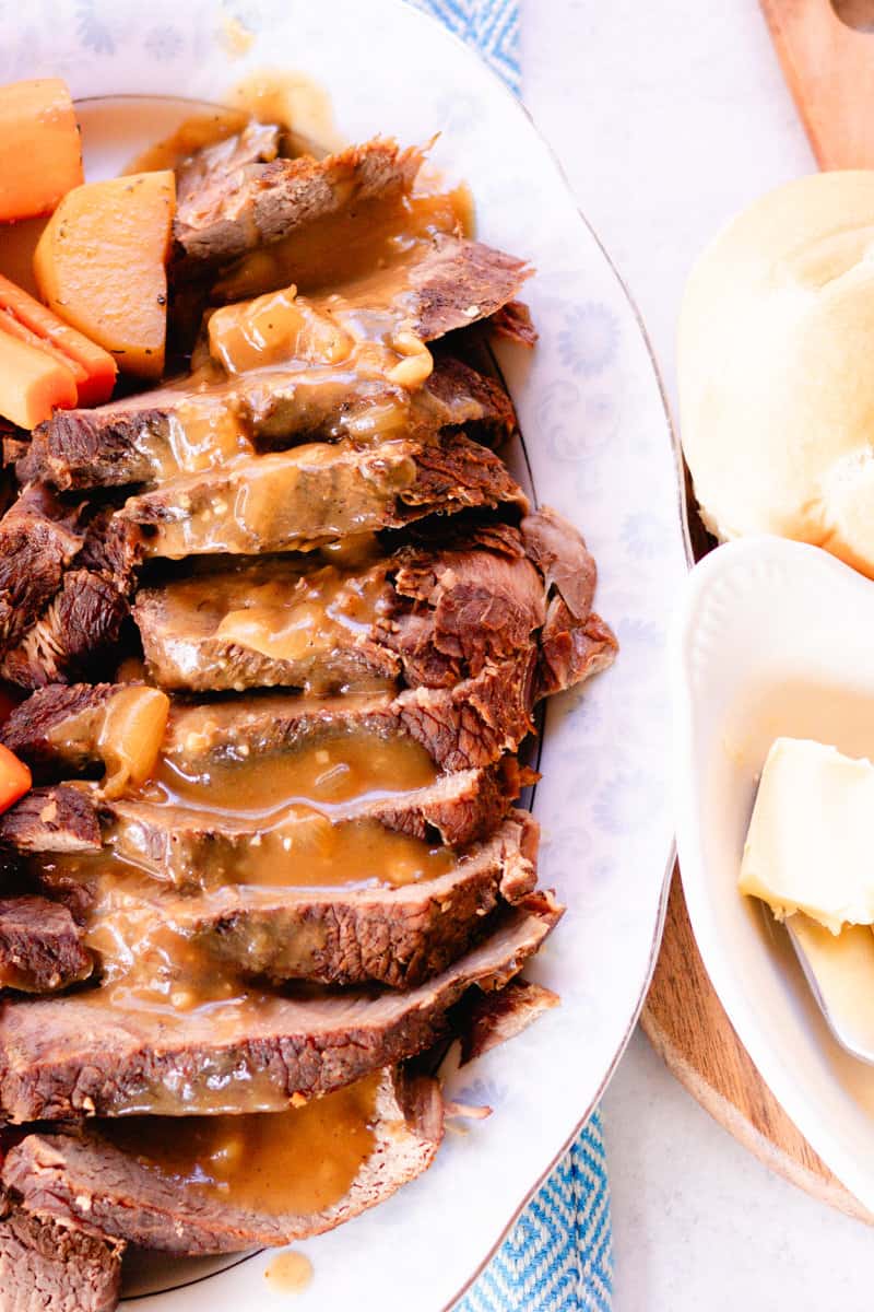 Sliced moose roast covered in gravy with butter and rolls on the side.