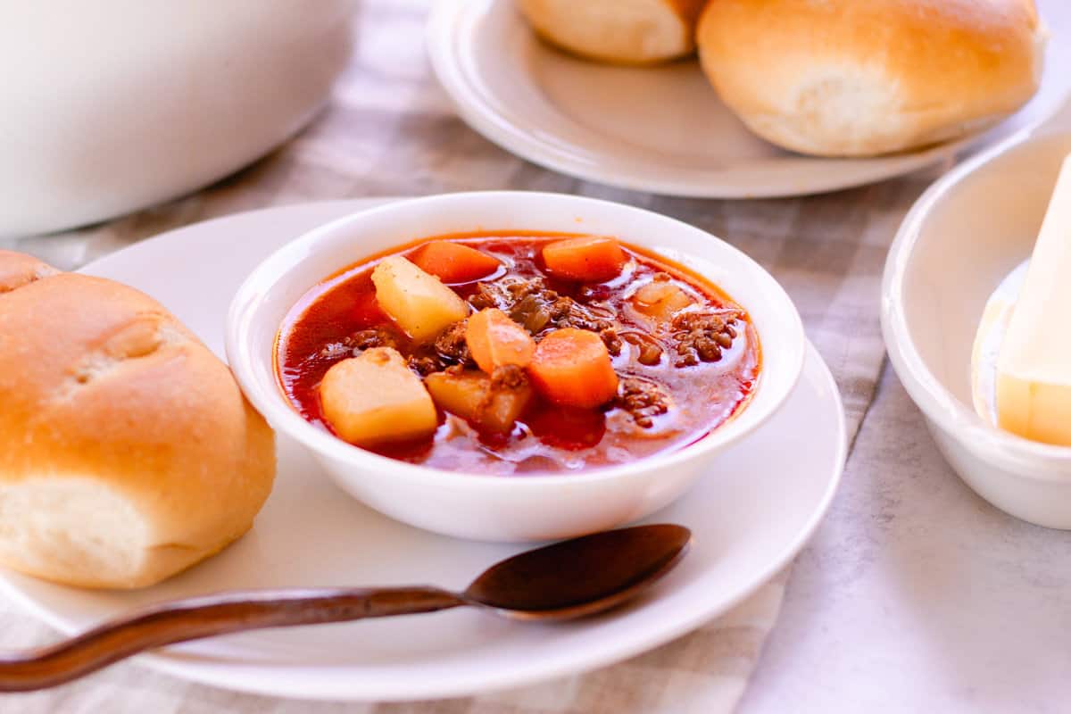 Hot bowl of moose meat soup with dinner rolls all on top of a blue checkered towel.