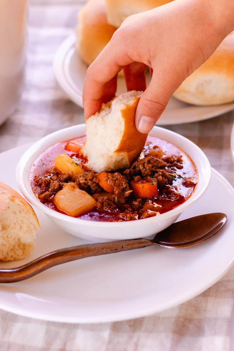 A small white bowl of hearty moose soup with a dinner roll and a golden spoon next to it.