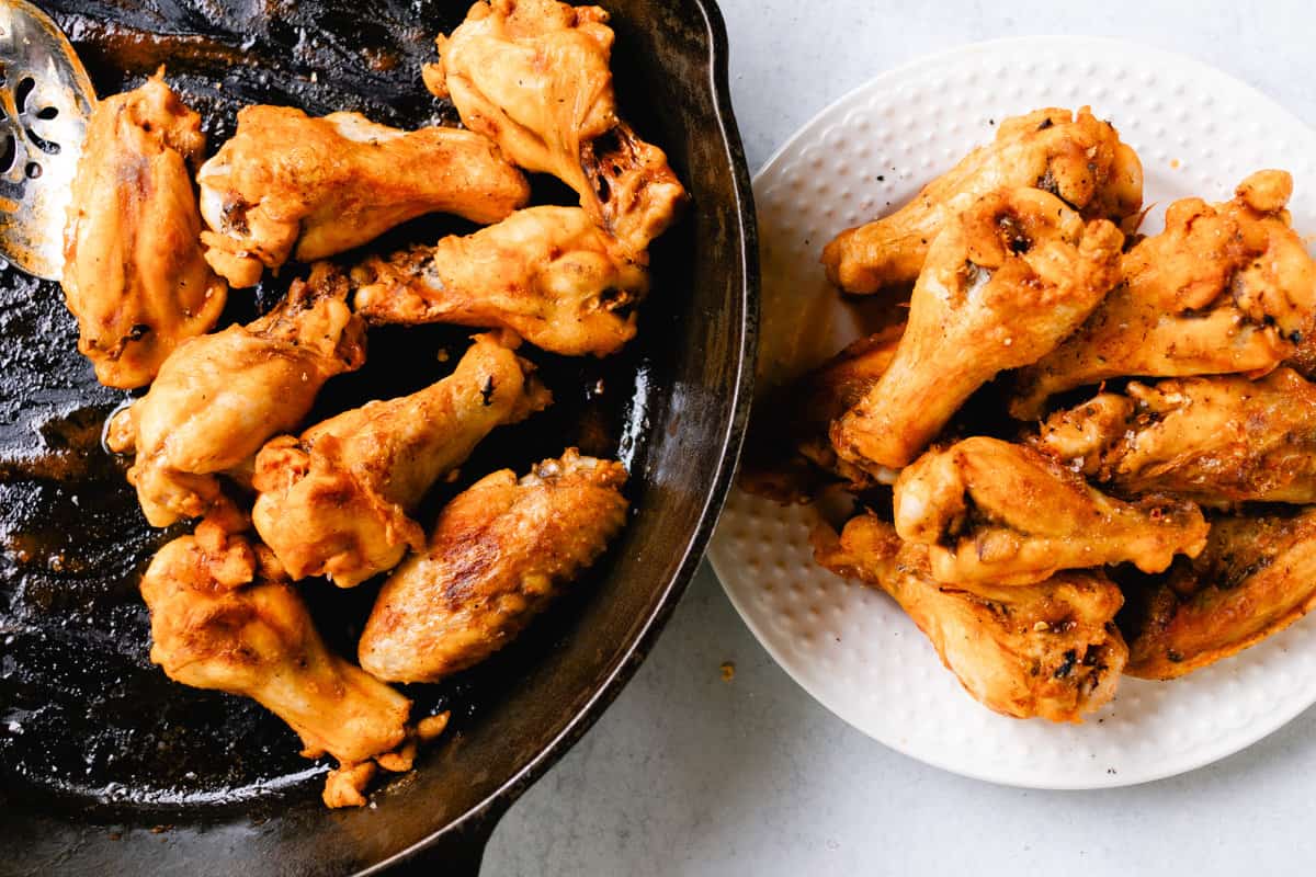 Transferring crispy chicken wings from a cast iron skillet onto a glass white plate.