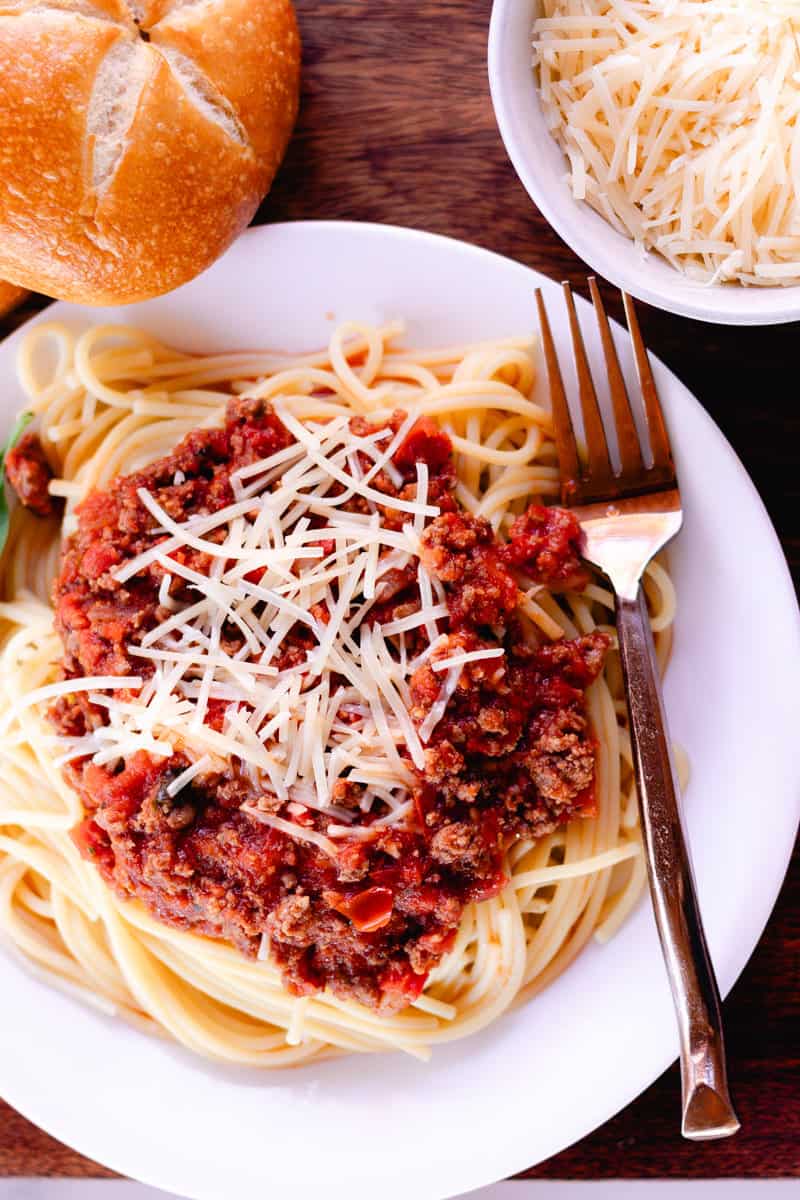 Deer meat pasta on a plate topped with shredded cheese and on the side is a roll and in a bowl is more cheese. 