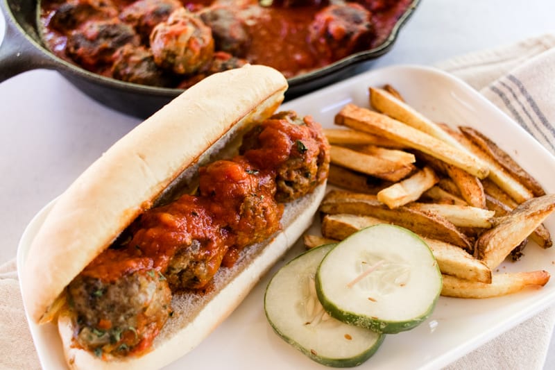 Meatballs on a roll with fries and pickles.