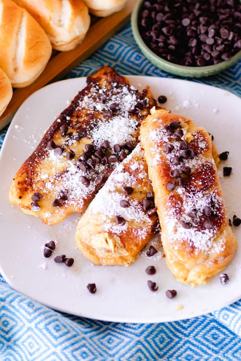 Crispy brioche toast topped with powered sugar and chocolate chips on a white plate with a bowl of chocolate chips in the corner. 