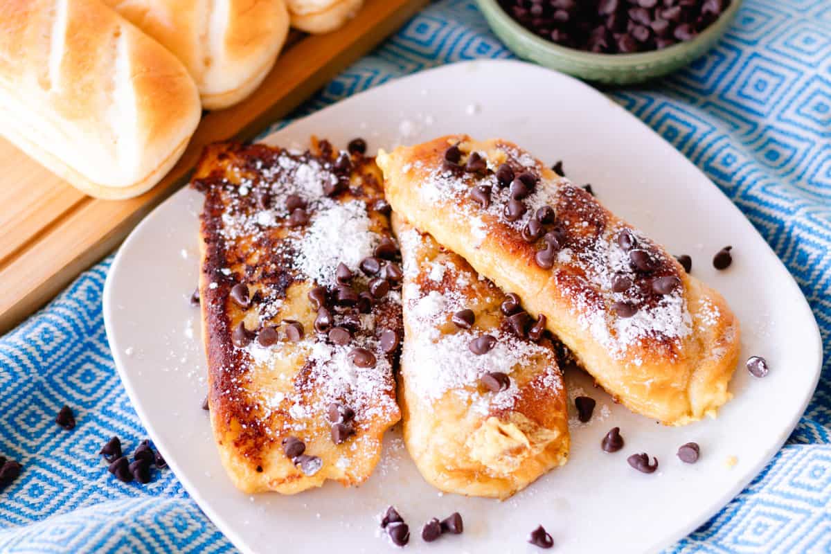 On a blue and white table cloth is a plate with three French toasts made from brioche bread topped with chocolate chips and powdered sugar.