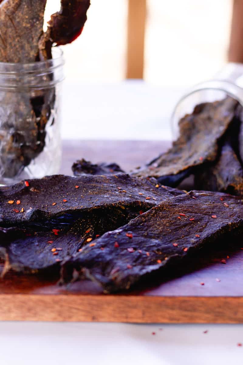 Fully cooked and dried elk jerky on a wooden drying board with several pieces in a glass jar in the background.