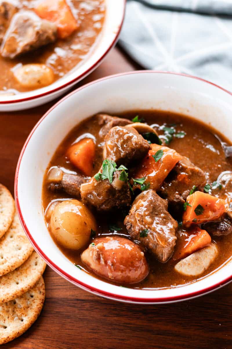 Thick hearty elk stew in a white bowl with a red rim on a dark wooden tabletop. There are also crackers in background and a blue and white tea towel.