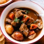 Hearty elk stew with thick chunks of elk meat, carrots and potatoes on a wooden table.