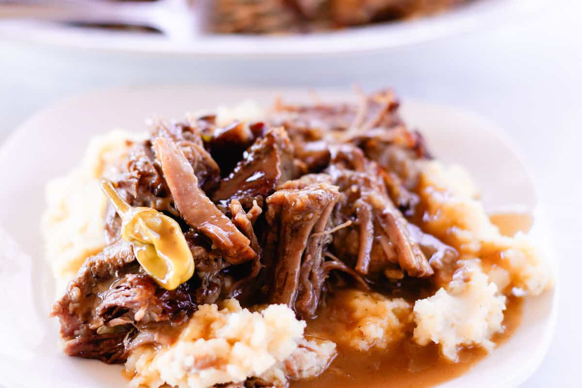 Slow cooked shredded pot roast overtop of mashed potatoes on a white plate.