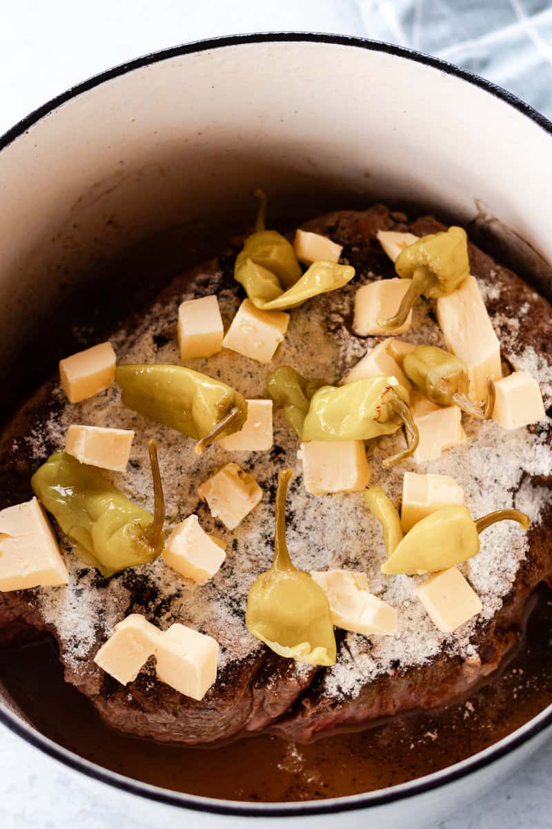 Butter, pepperoncini's, and seasoning on top of a pot roast in a dutch oven.