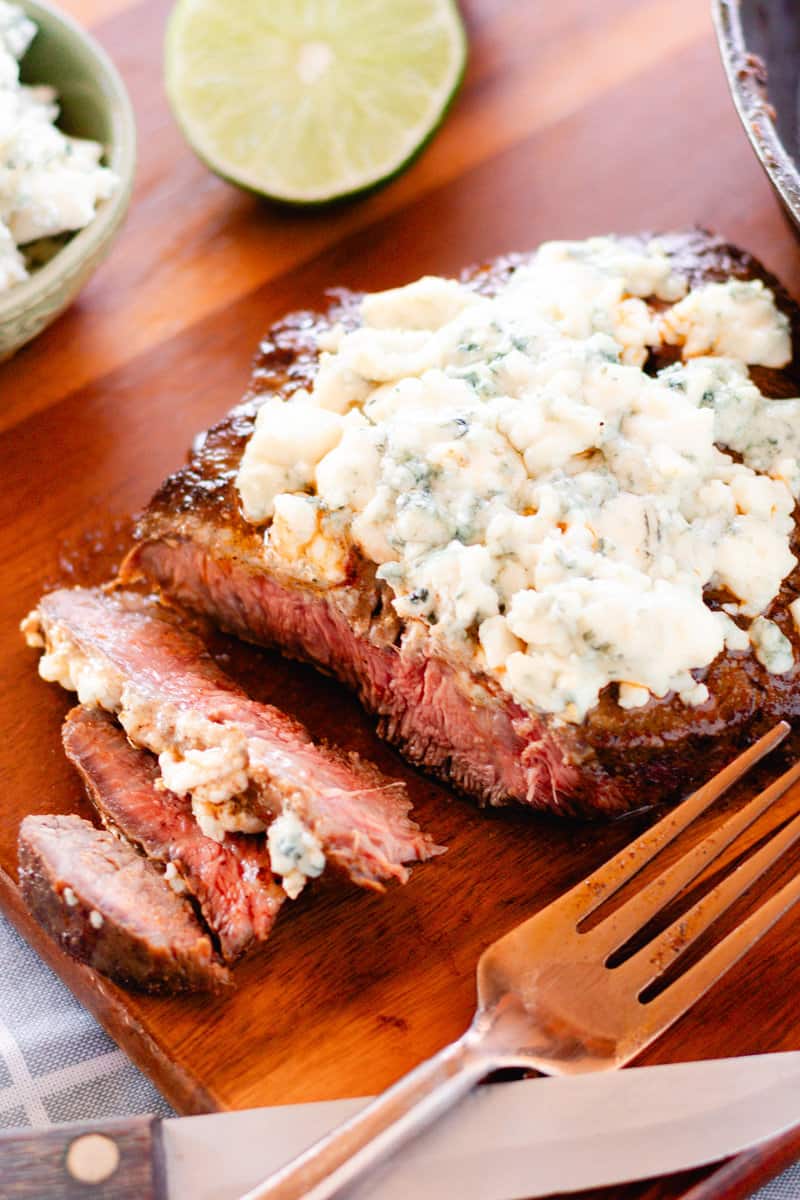 Flat iron steak with blue cheese crumbles on a wooden serving tray with a fork and lime slice.