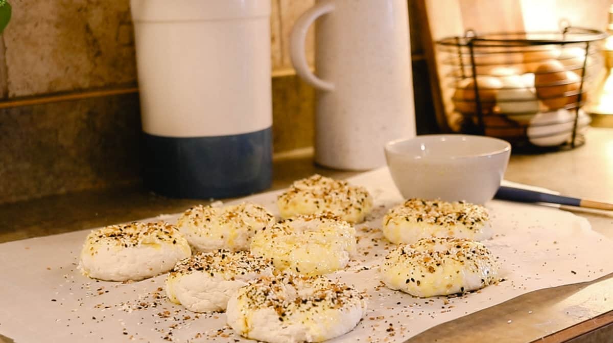 Raw, shaped bagels on a sheet of parchment paper on a kitchen counter that are brushed with egg wash and sprinkled with 