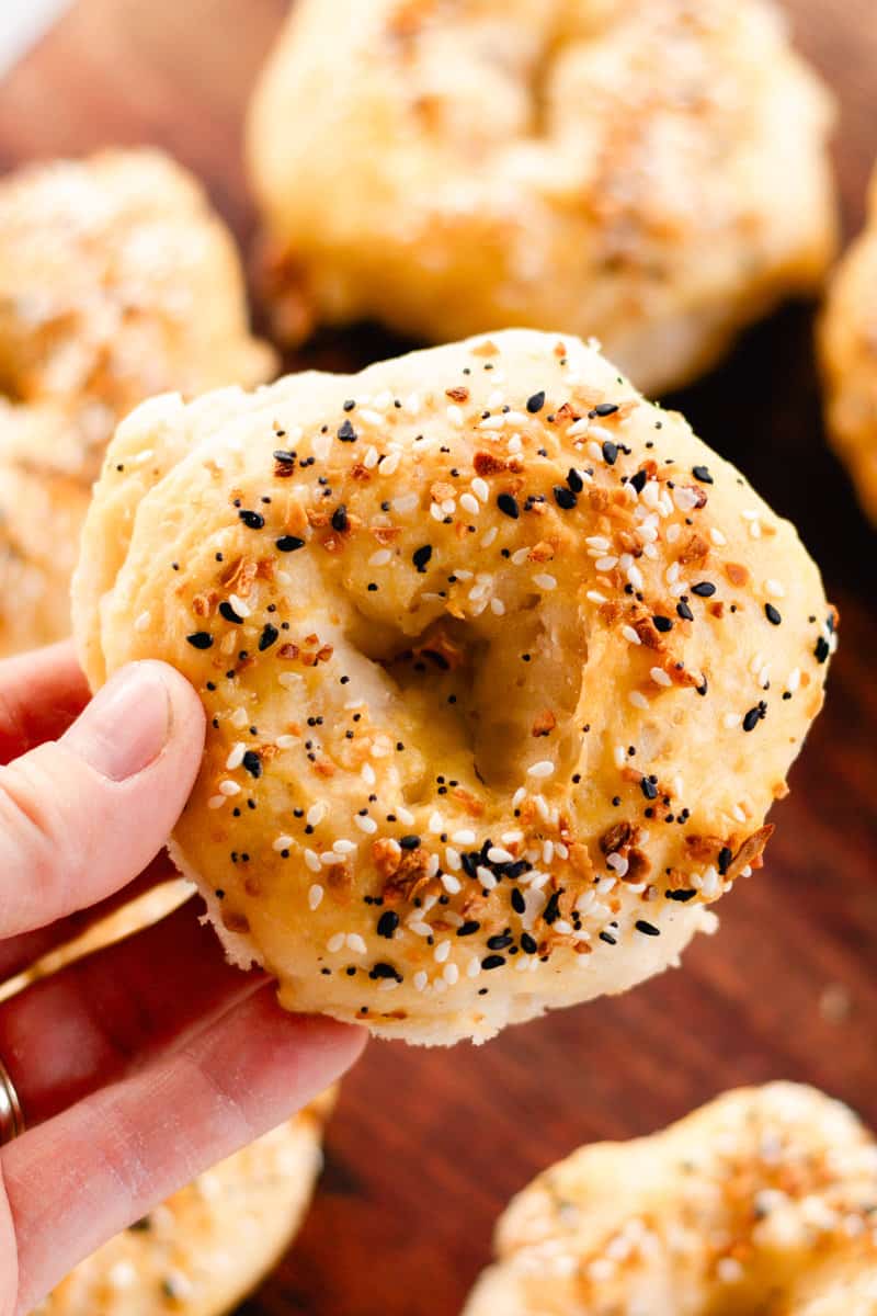 A woman's hand holding a homemade Greek yogurt bagel covered in everything bagel seasoning with more bagels in the background.