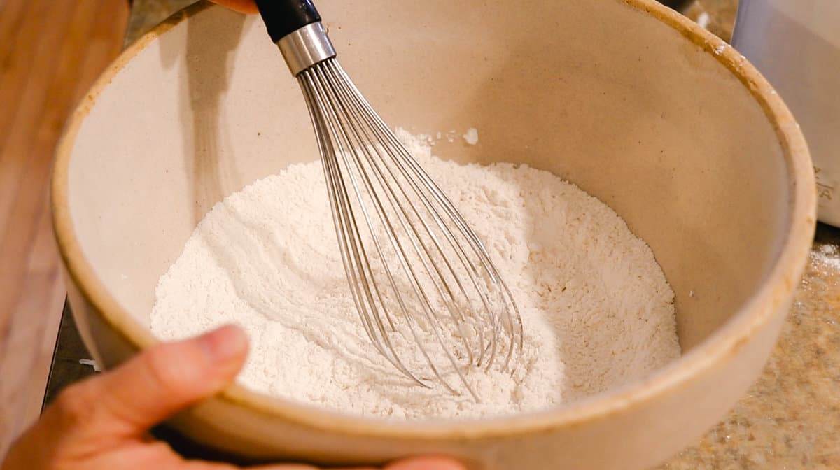 A woman whisking flour, baking powder, and salt in a stoneware mixing bowl.