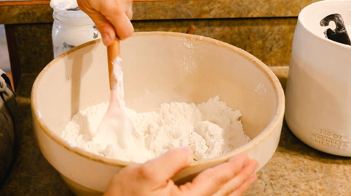 A woman using a spatula to mix Greek yogurt and the dry ingredients for homemade bagels in a large stoneware mixing bowl.