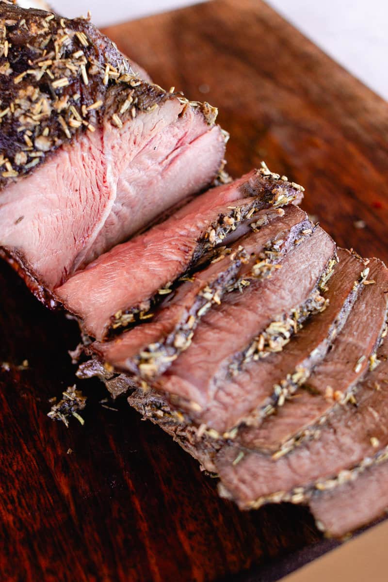 An eye of round roast with an olive oil and herb coating on a wooden cutting board sliced into thin pieces.