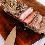 Thinly sliced eye of round roast with a rosemary and thyme marinade over it on a wooden cutting board.