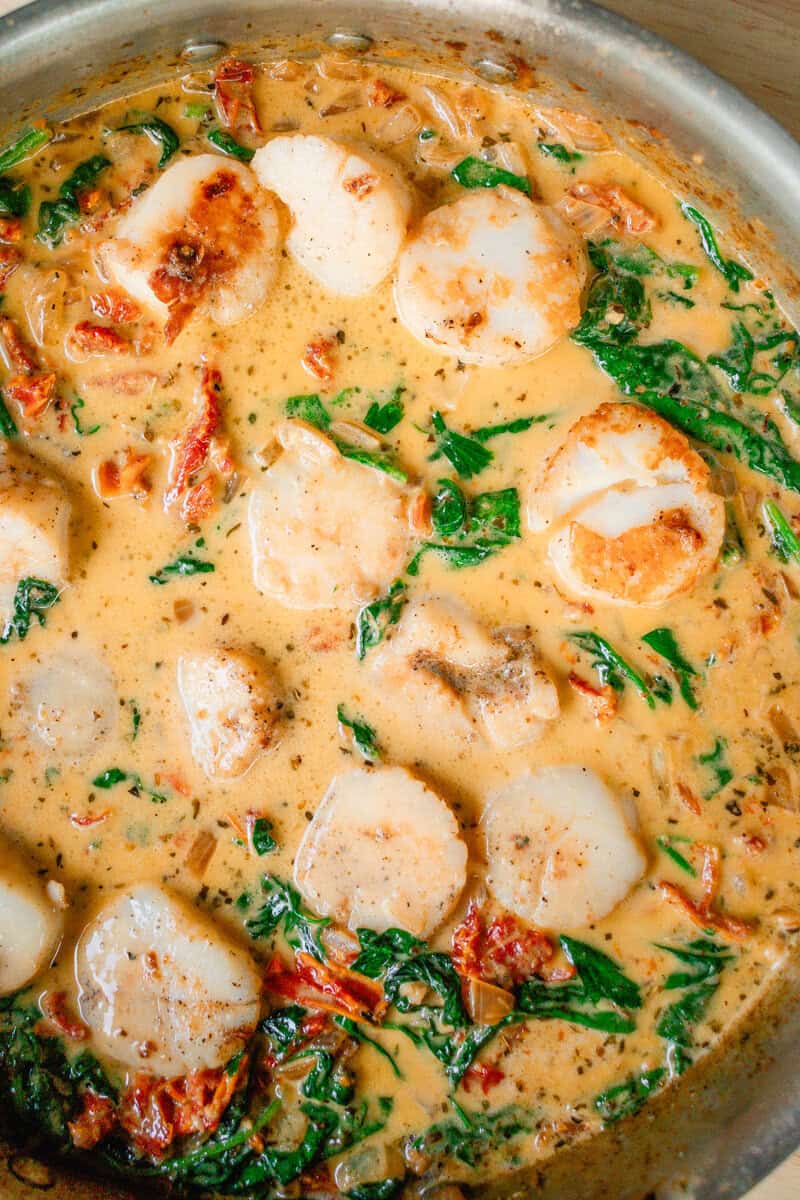 Several sea scallops simmering in a frying pan of creamy sauce.