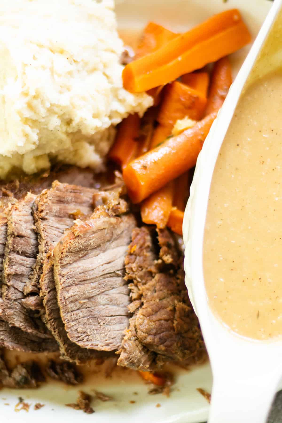 A rump roast sliced thin and stacked on a serving plate with mashed potatoes, carrots and gravy.