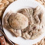 Boiled pork chops with spices served on a white plate with a fork and steak knife on a raffia trivet.