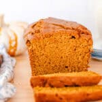 Sliced pumpkin bread on a wooden table with a stack of plates and a tea pot in the background.