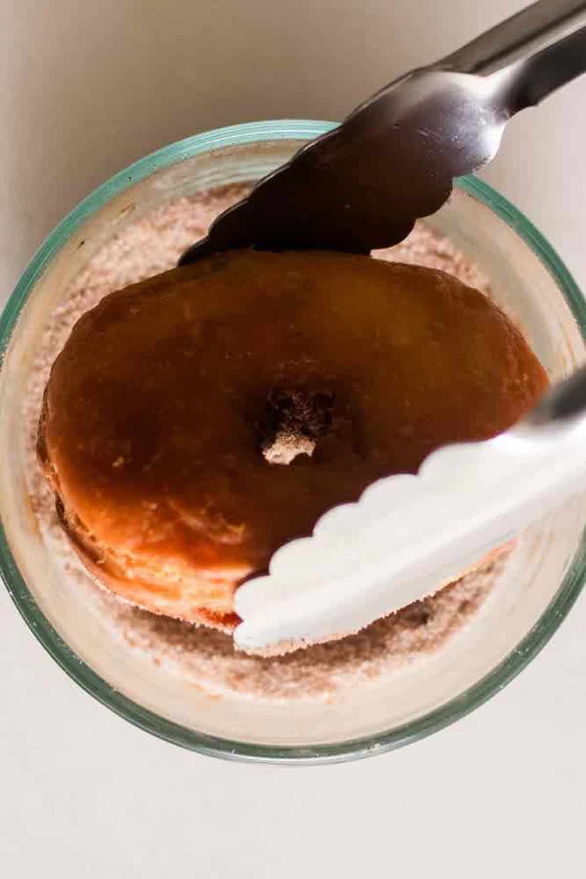 Using tongs to dip the hot donut in a cinnamon sugar mixture. 