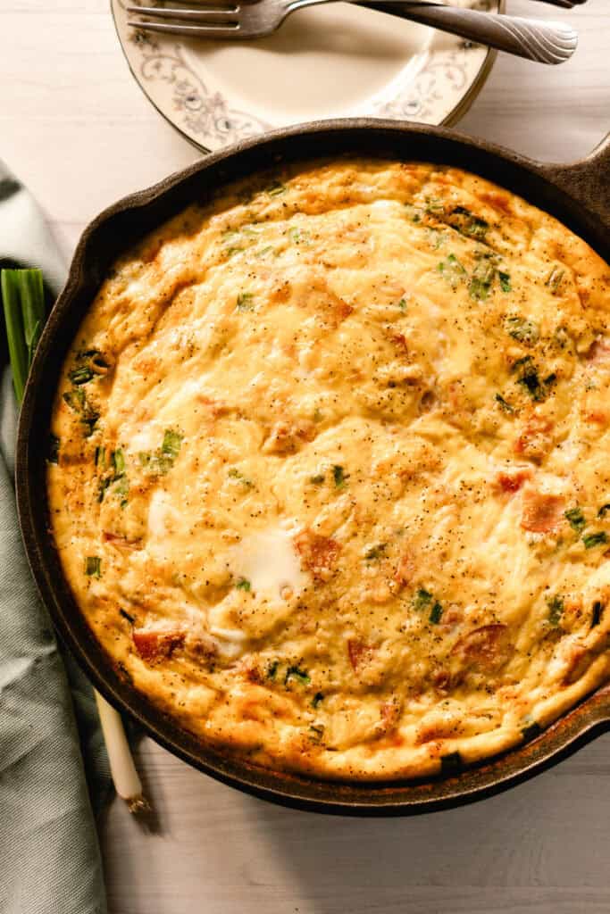 Cast iron quiche with hunks of ham on a white wooden table with a blue towel next to it.