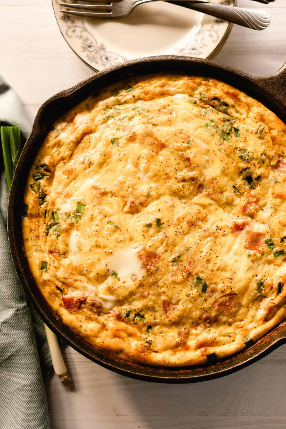 Cast iron quiche with hunks of ham on a white wooden table with a blue towel next to it.