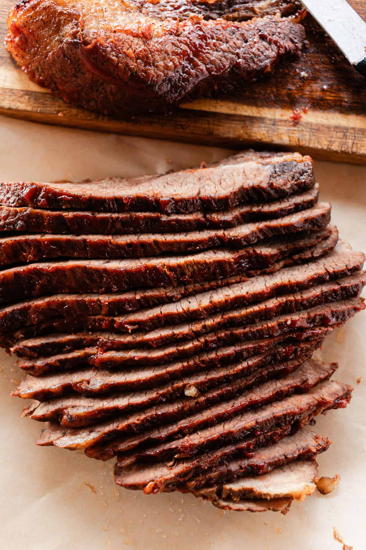 Thin sliced beef brisket on parchment paper with a big hunk of it sitting in the background on a wooden cutting board. 