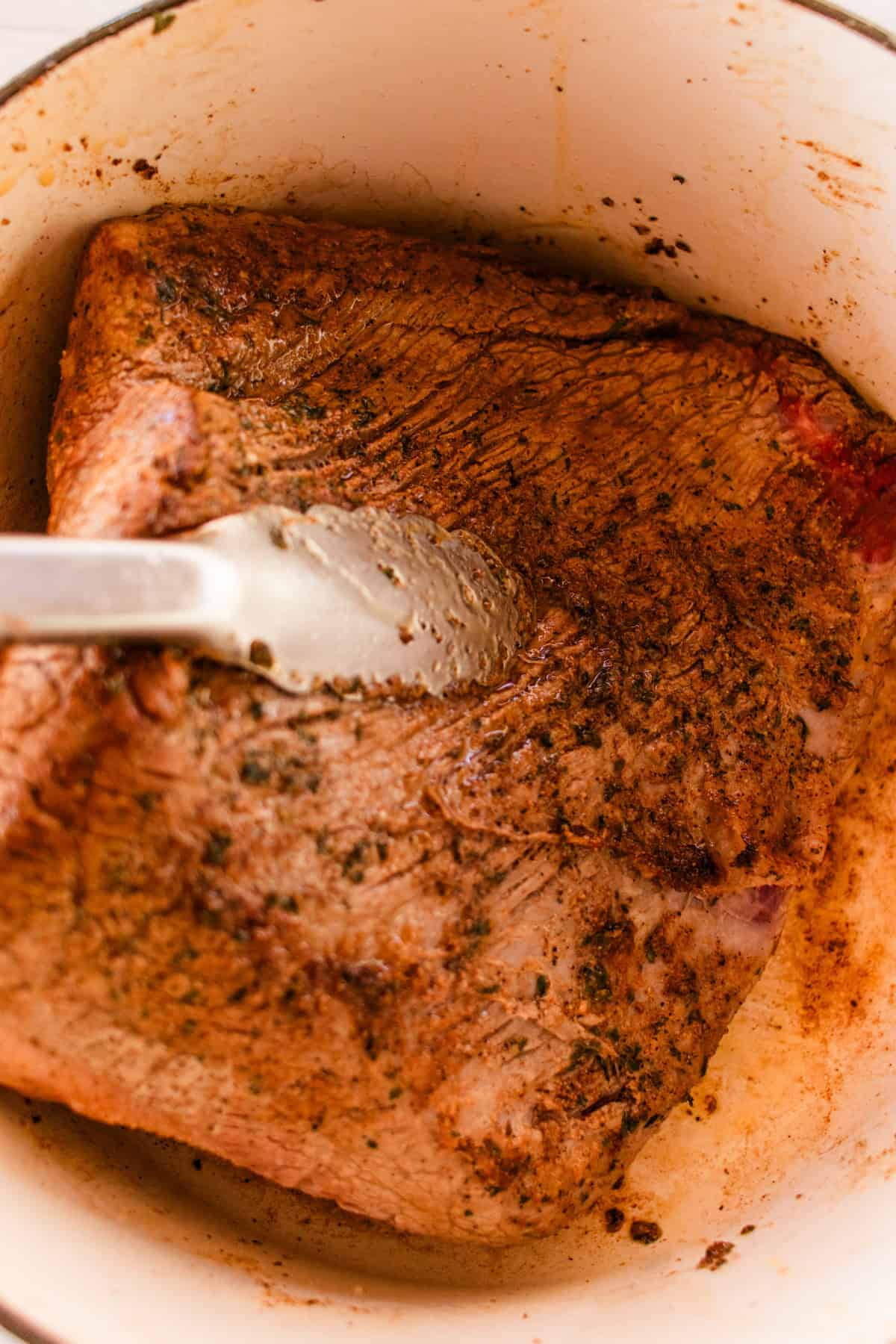 The brisket searing in a dutch oven while tongs are being used to flip it over. 