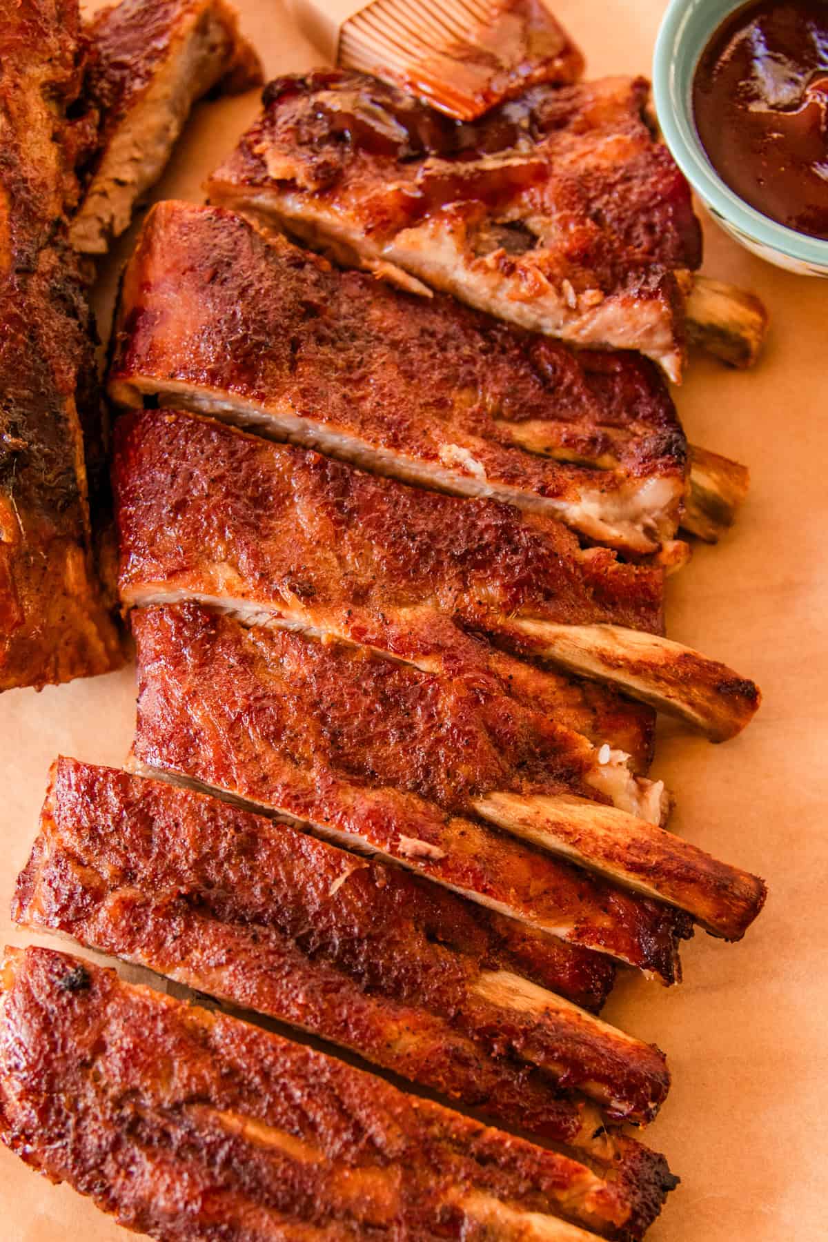 Spare ribs sliced and resting on parchment paper with a small bowl of bbq sauce next to it.
