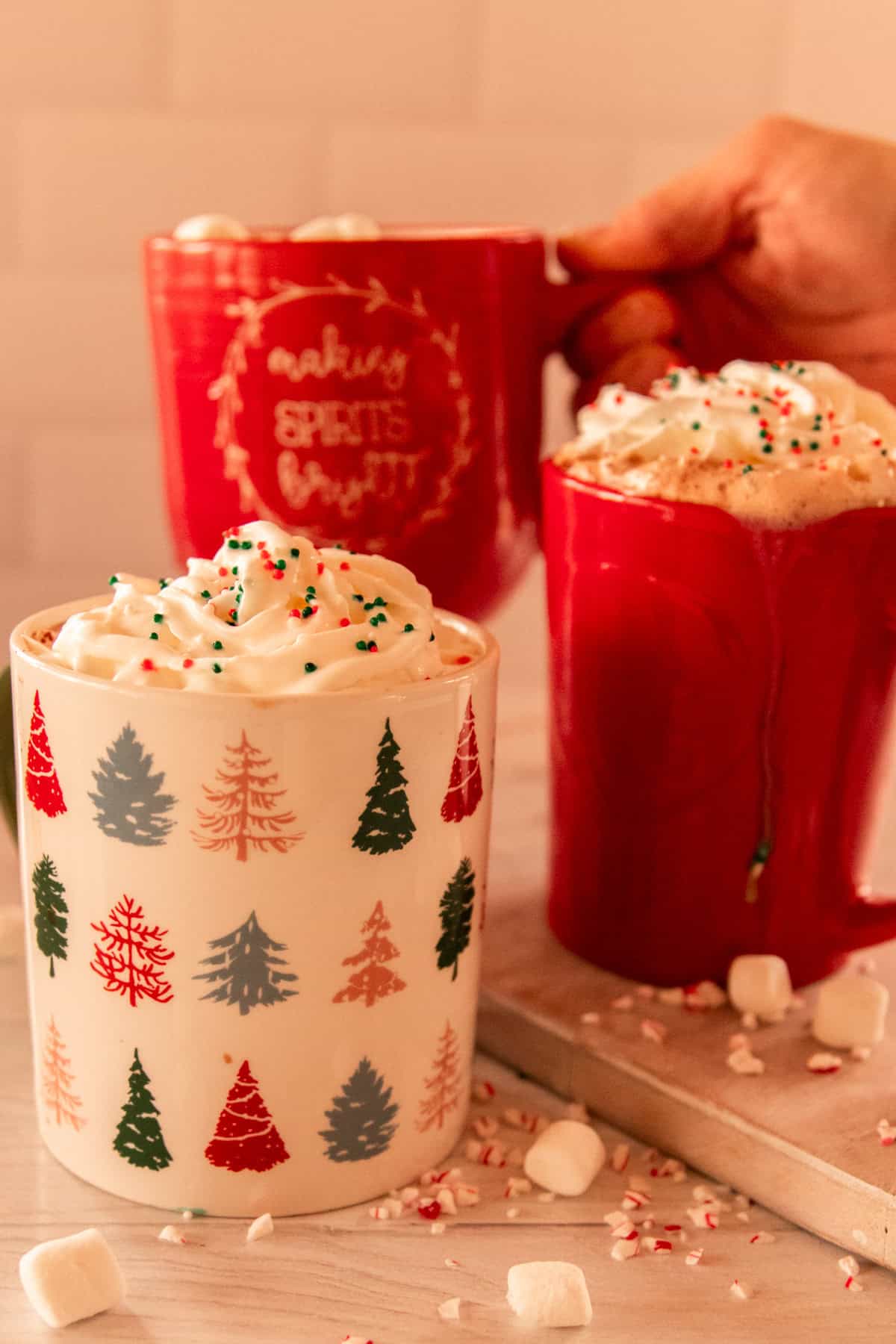 Homemade hot chocolate in 3 different holiday mugs on a white table with crushed candy canes spread around them. 