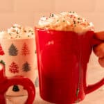 Holding a red mug with homemade hot chocolate and whipped cream overflowing the top with fun sprinkles.