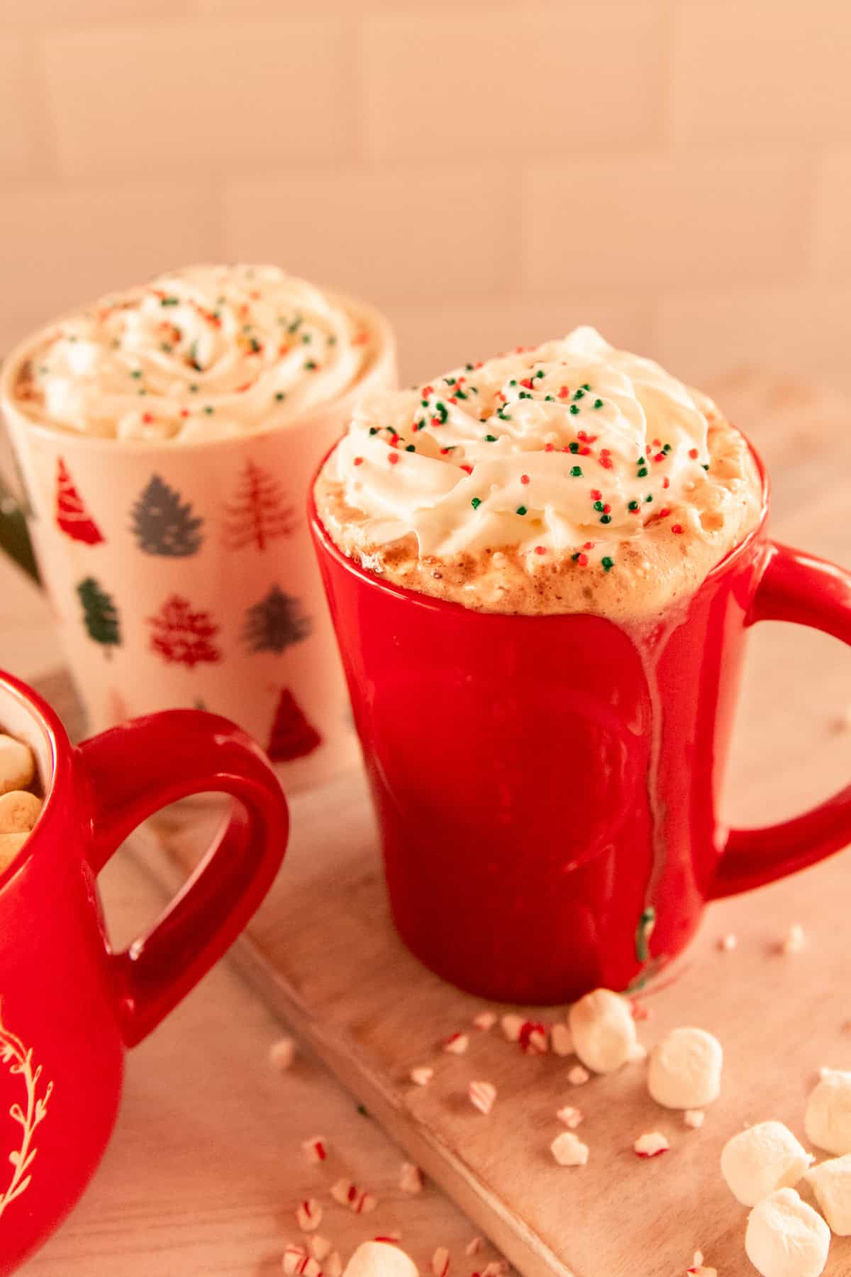 3 mugs with homemade hot cocoa with whipped cream, sprinkles and marshmallows around them. 