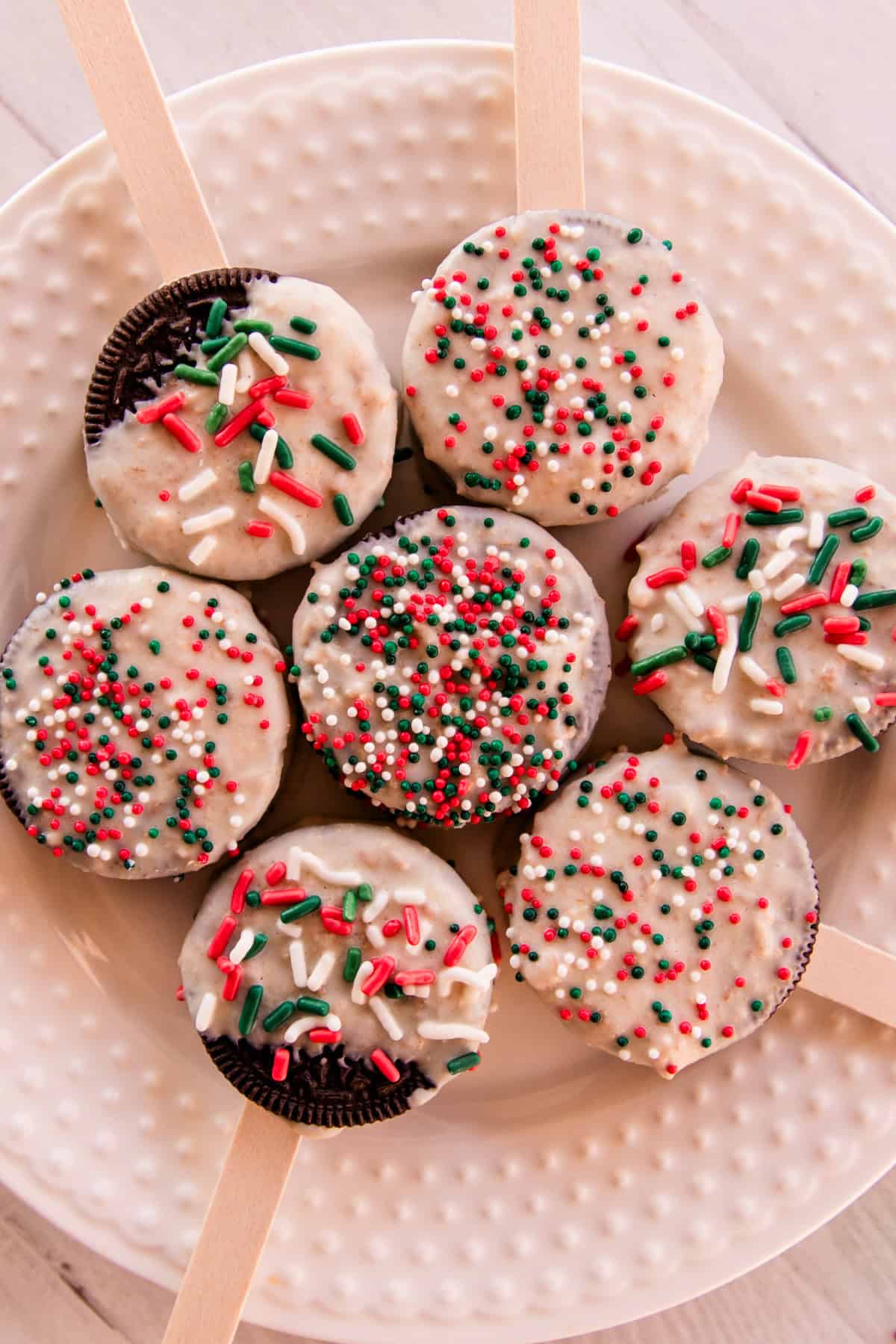7 Christmas decorated Oreos that are dipped in white chocolate and topped with sprinkles on a white plate. 
