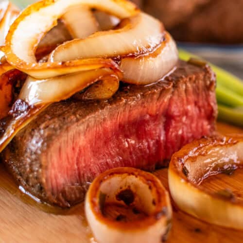 Medium rare venison steak, sliced on a cutting board with sautéed onions piled on top.