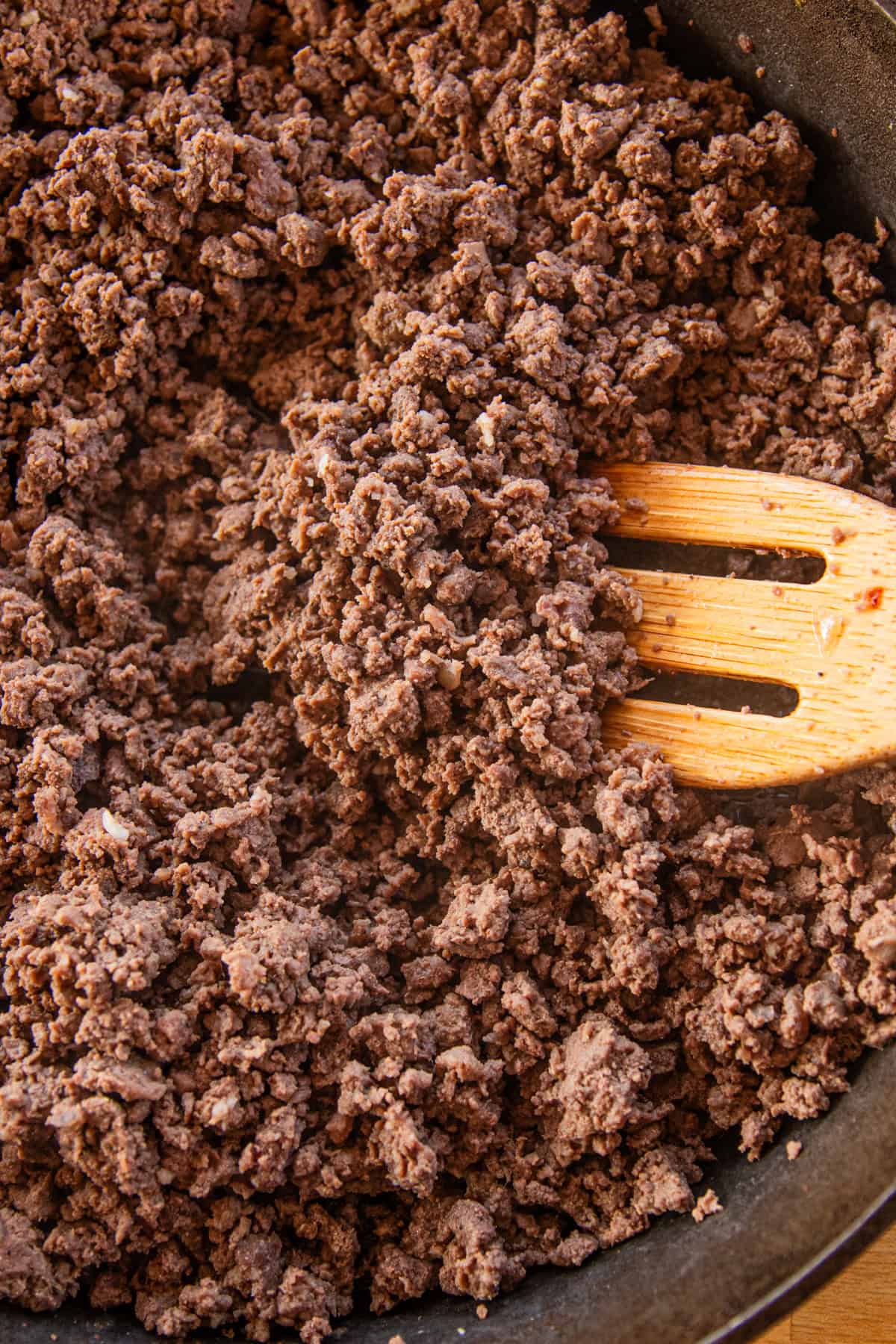 Browned ground beef in a cast iron skillet, being stirred with a wooden spatula. 