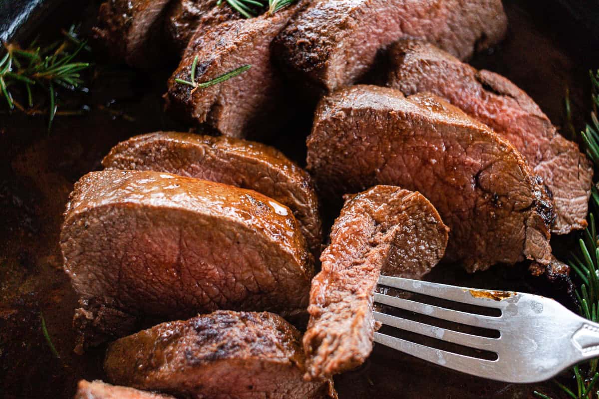 Thin sliced venison tenderloin with a large piece of butter melting on top of it and a few sprigs of rosemary in the skillet next to it.