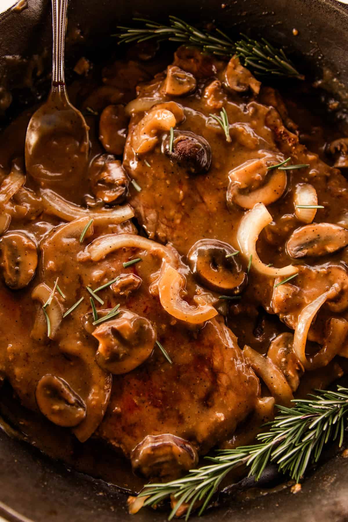 Spooning gravy over top of the round steaks that are simmering in a large cast iron skillet.