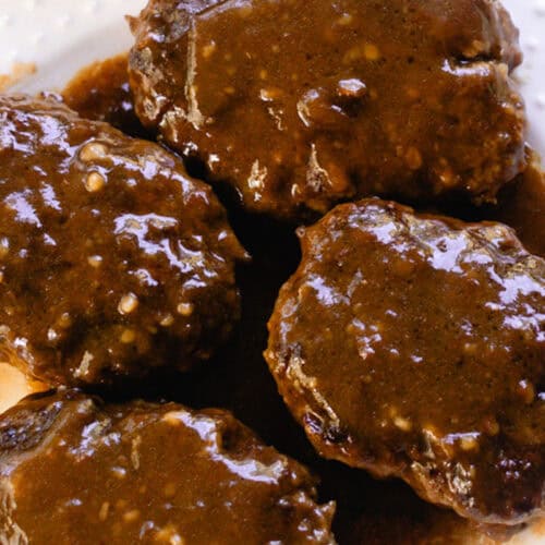 4 salisbury steak patties on a white serving plate with dark brown gravy on top.