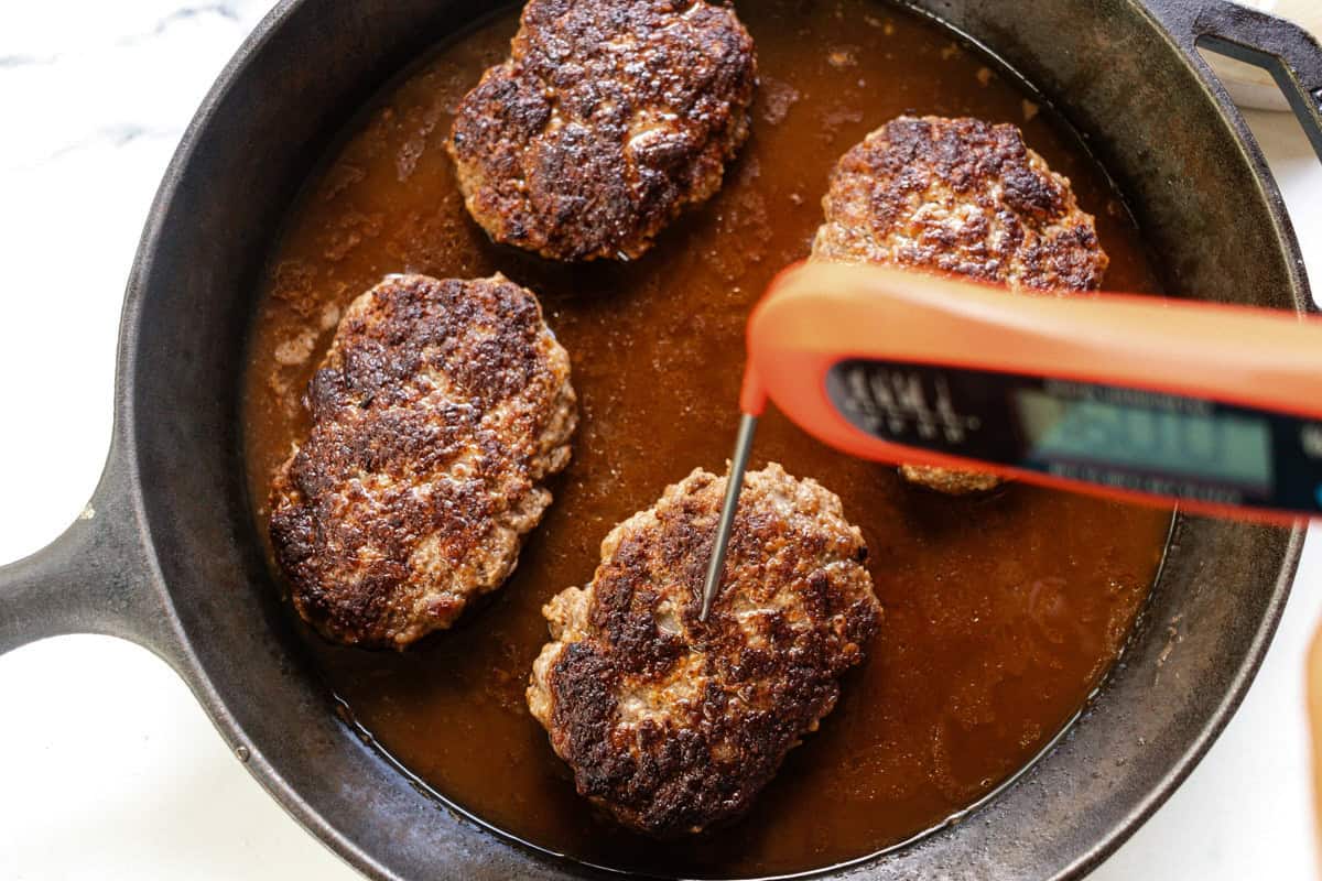 Probing one of the salisbury steaks in the skillet with a meat thermometer to check the internal temperature.